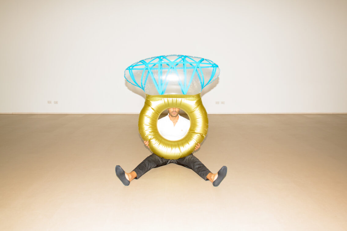 A man sitting on the floor during a workday pause, with a large golden and blue inflatable structure encircling his upper body, creating an artistic and surreal scene