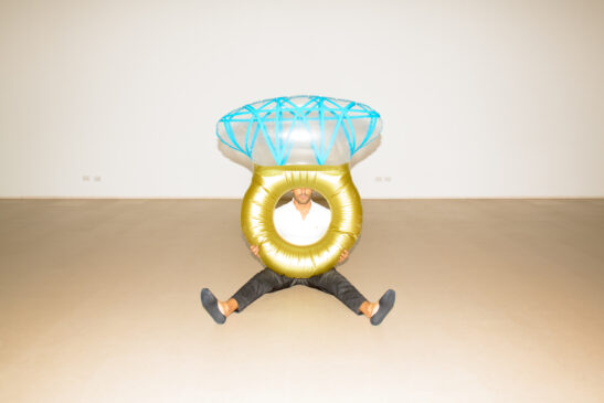 A man sitting on the floor during a workday pause, with a large golden and blue inflatable structure encircling his upper body, creating an artistic and surreal scene